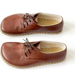 Peut inclure: Deux chaussures en cuir marron avec lacets, sur fond blanc. Les chaussures ont un bout arrondi et une semelle cousue. Le cuir est lisse et les lacets sont marron. Chaque chaussure mesure environ 27 cm de long.