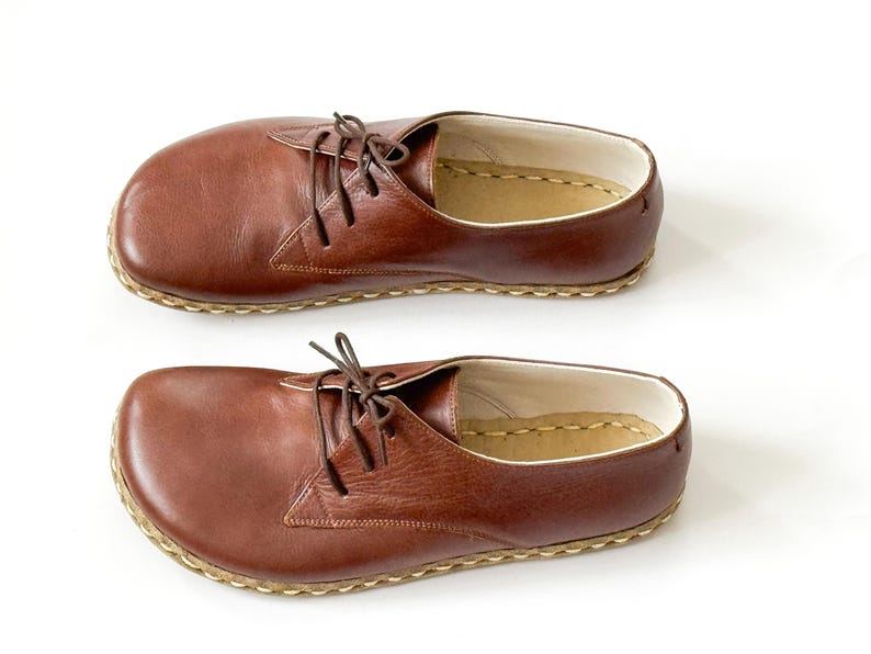 Peut inclure: Deux chaussures en cuir marron avec lacets, sur fond blanc. Les chaussures ont un bout arrondi et une semelle cousue. Le cuir est lisse et les lacets sont marron. Chaque chaussure mesure environ 27 cm de long.