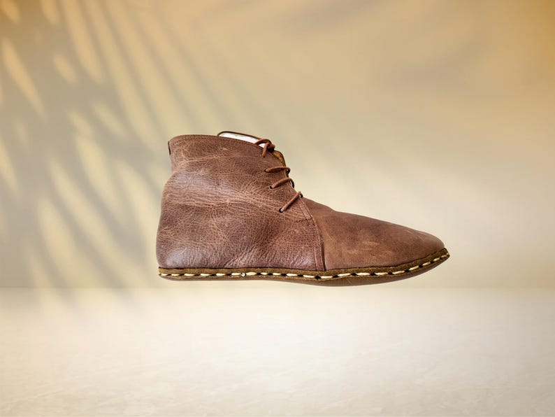 Puede incluir: Una bota de cuero marr&oacute;n con cordones. La bota tiene una superficie texturizada y una suela cosida. El fondo es beige neutro con la sombra de una hoja de palma.