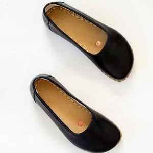 May include: A pair of black leather ballet flats. The shoes have a rounded toe, a light brown insole, and a copper-coloured rivet on the side. The soles are a light brown colour with visible stitching.