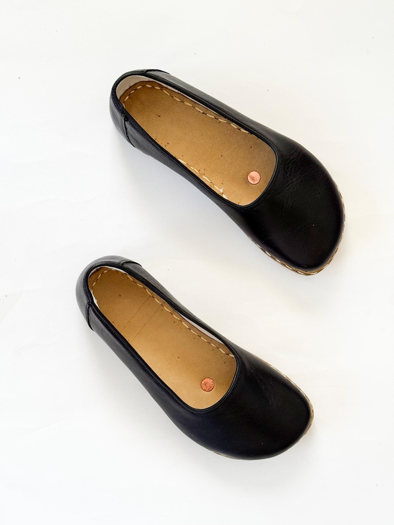 May include: A pair of black leather ballet flats. The shoes have a rounded toe, a light brown insole, and a copper-coloured rivet on the side. The soles are a light brown colour with visible stitching.