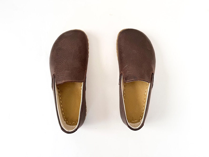 Pu&ograve; includere: Un paio di scarpe slip-on in pelle marrone scuro. Le scarpe hanno la punta arrotondata e l'interno marrone chiaro. Le suole sono di colore marrone chiaro. Le scarpe sono su uno sfondo bianco.