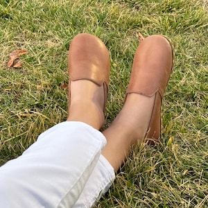 Pode incluir: Um par de sapatos slip-on de couro castanho claro. Os sapatos est&atilde;o nos p&eacute;s de uma pessoa, com cal&ccedil;as brancas vis&iacute;veis. Os sapatos est&atilde;o na relva verde.