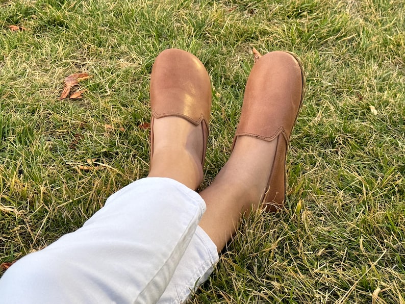 Pode incluir: Um par de sapatos slip-on de couro castanho claro. Os sapatos est&atilde;o nos p&eacute;s de uma pessoa, com cal&ccedil;as brancas vis&iacute;veis. Os sapatos est&atilde;o na relva verde.