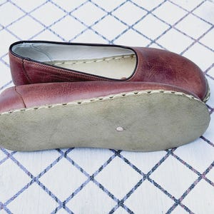 May include: A pair of burgundy leather slip-on shoes. The shoes have a rounded toe and a stitched sole. The interior is a light color, and the sole is a light gray. The shoes are displayed on a white and blue checkered surface.