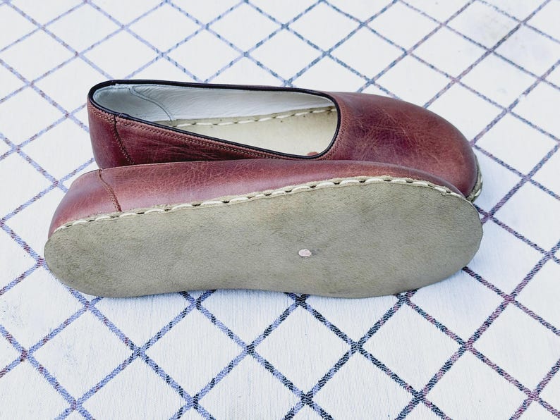 May include: A pair of burgundy leather slip-on shoes. The shoes have a rounded toe and a stitched sole. The interior is a light color, and the sole is a light gray. The shoes are displayed on a white and blue checkered surface.