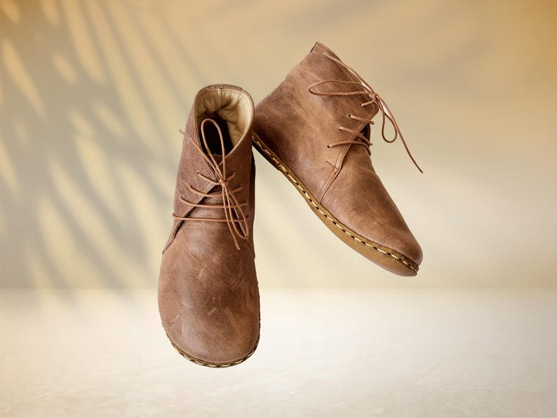 Puede incluir: Un par de botines de cuero marr&oacute;n con cordones. Las botas tienen un aspecto texturizado y una suela cosida. El fondo es de un color suave y neutro con una sombra sutil.