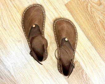 May include: A pair of brown leather sandals with white stitching and ankle straps. The sandals have a minimalist design with a round wooden button detail. The shoes are displayed on a light-colored wooden surface.