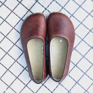 May include: A pair of brown leather slip-on shoes. The shoes have a rounded toe and a light-colored interior. The shoes are placed on a white surface with a blue and gray diamond pattern.