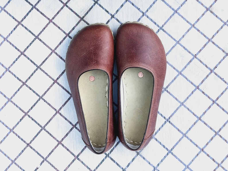 May include: A pair of brown leather slip-on shoes. The shoes have a rounded toe and a light-colored interior. The shoes are placed on a white surface with a blue and gray diamond pattern.