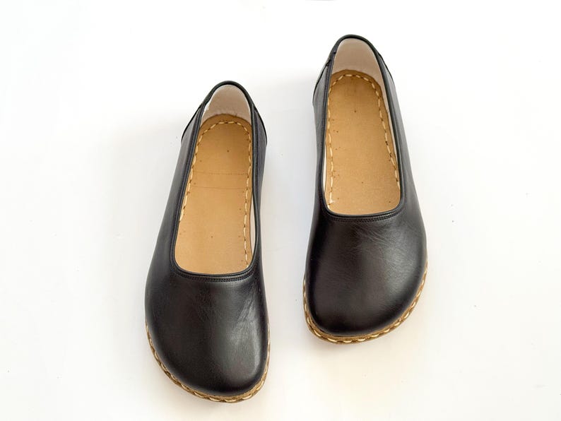 May include: A pair of black leather flats with a rounded toe and a tan insole. The shoes have a stitched sole and a simple, classic design. The shoes are likely for everyday wear.