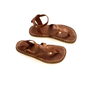 Op de afbeelding: Een paar bruin lederen sandalen met een teenbandje en enkelbandje. De sandalen hebben een gestikte zool en een gespsluiting. Het leer heeft een getextureerde uitstraling.