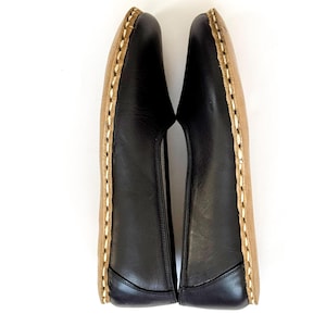May include: A pair of black leather slip-on shoes with tan soles. The shoes have a stitched design around the edges. The shoes are displayed on a white background.