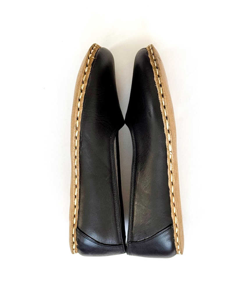 May include: A pair of black leather slip-on shoes with tan soles. The shoes have a stitched design around the edges. The shoes are displayed on a white background.