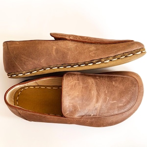 Peut inclure: Paire de mocassins en cuir marron. Ces chaussures à enfiler présentent un design classique avec un bout arrondi et des coutures blanches visibles autour de la semelle. Le cuir a un aspect vieilli, donnant une sensation vintage.