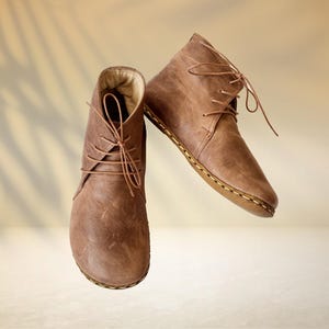 Puede incluir: Un par de botines de cuero marr&oacute;n con cordones. Las botas tienen un aspecto texturizado y una suela cosida. El fondo es de un color suave y neutro con una sombra sutil.