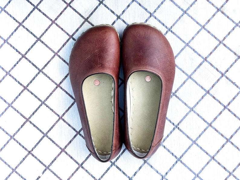 May include: A pair of brown leather clogs, viewed from above, resting on a white and blue checkered surface. The clogs have a rounded toe and a light-colored interior. Each clog has a small, circular detail near the heel.