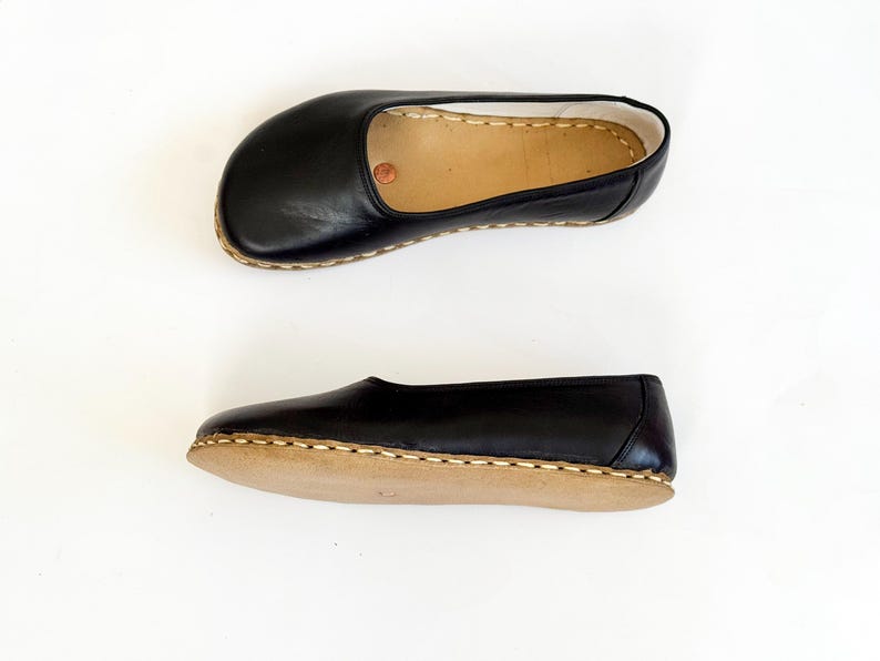 May include: Two black leather slippers with a rounded toe and a tan sole are shown against a white backdrop. The slippers have a simple design with visible stitching around the sole. The leather appears smooth and the overall style is minimalist.