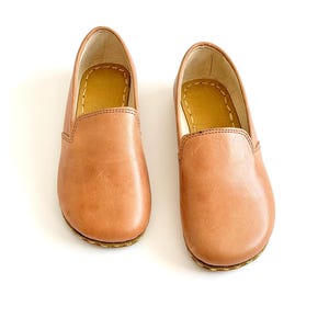 Pode incluir: Um par de sapatos slip-on de couro castanho claro. Os sapatos t&ecirc;m bico arredondado e sola cosida. O interior &eacute; de cor castanho claro. Os sapatos est&atilde;o sobre um fundo branco.