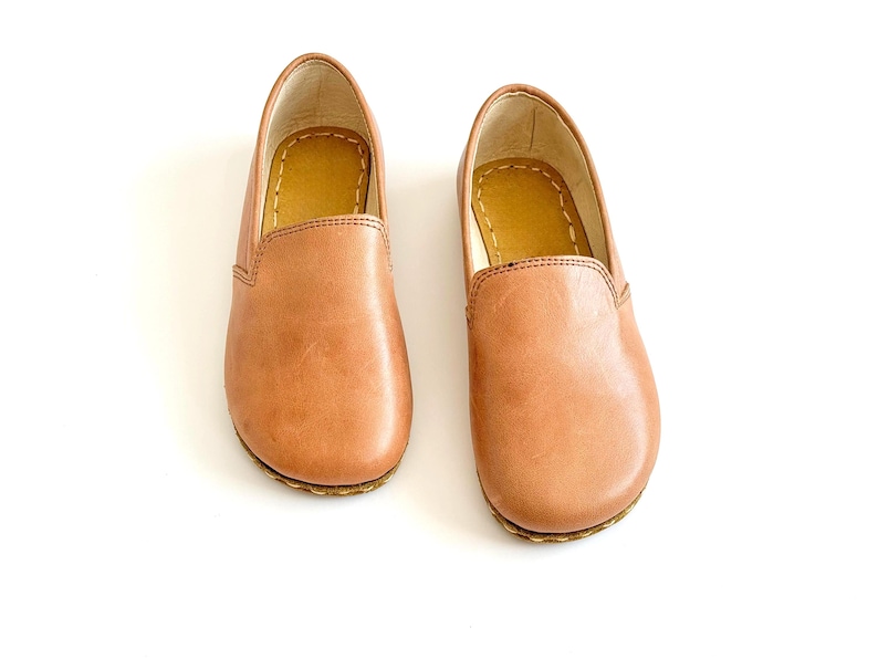 Pode incluir: Um par de sapatos slip-on de couro castanho claro. Os sapatos t&ecirc;m bico arredondado e sola cosida. O interior &eacute; de cor castanho claro. Os sapatos est&atilde;o sobre um fundo branco.