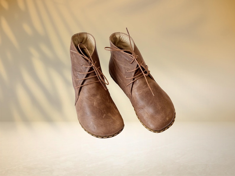 Puede incluir: Un par de botines de cuero marr&oacute;n con cordones. Los botines tienen una punta redondeada y un dise&ntilde;o texturizado en el lateral. Las suelas son de color marr&oacute;n claro. El fondo es un degradado de beige y bronceado.