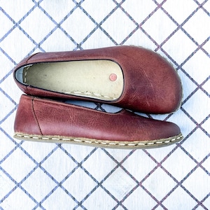 May include: A pair of burgundy leather slip-on shoes. The shoes are flat with a rounded toe and a stitched sole. The shoes are displayed on a white surface with a blue diamond pattern.