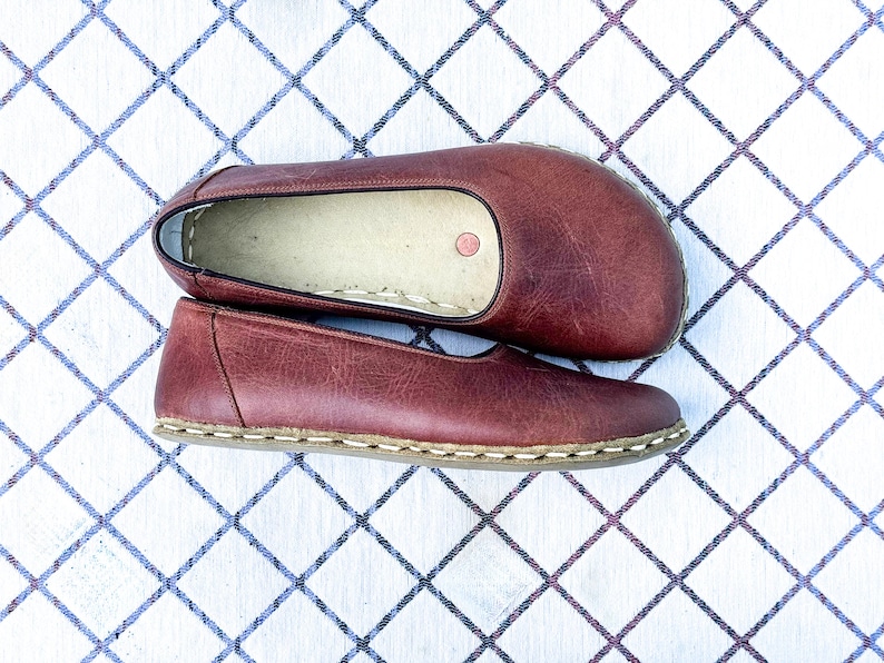 May include: A pair of burgundy leather slip-on shoes. The shoes are flat with a rounded toe and a stitched sole. The shoes are displayed on a white surface with a blue diamond pattern.