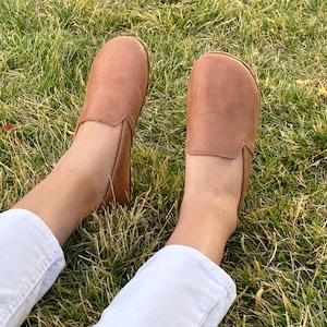 Pode incluir: Um par de sapatos slip-on de couro castanho claro. Os sapatos est&atilde;o sobre uma superf&iacute;cie gramada. Os sapatos t&ecirc;m uma biqueira arredondada e um perfil baixo. Os sapatos s&atilde;o de cor neutra.