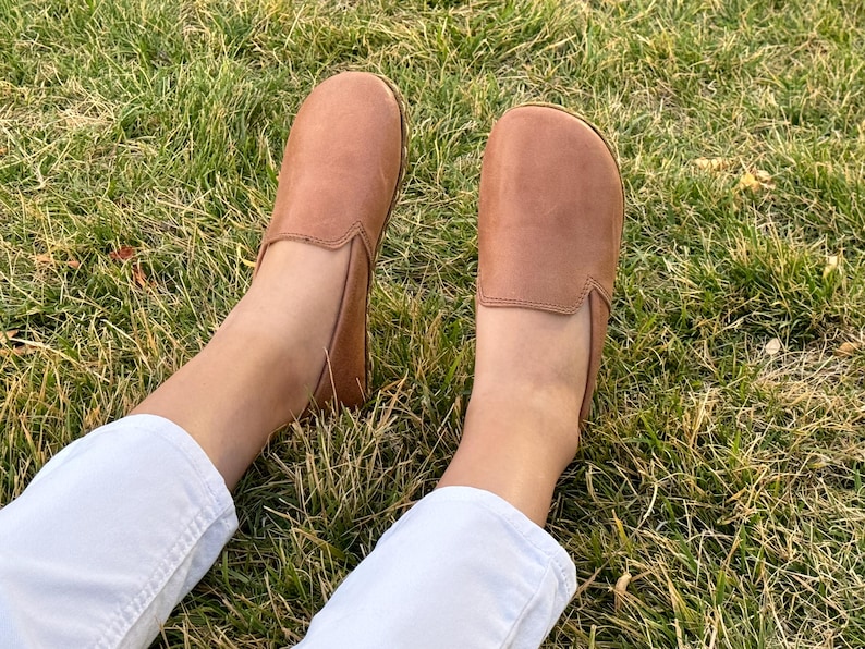 Pode incluir: Um par de sapatos slip-on de couro castanho claro. Os sapatos est&atilde;o sobre uma superf&iacute;cie gramada. Os sapatos t&ecirc;m uma biqueira arredondada e um perfil baixo. Os sapatos s&atilde;o de cor neutra.