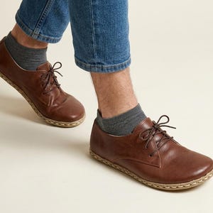 Peut inclure: Chaussures &agrave; lacets en cuir marron avec une semelle cousue. Les chaussures sont port&eacute;es avec des chaussettes grises et un jean bleu. Les chaussures ont un design classique et une esth&eacute;tique naturelle et terreuse.
