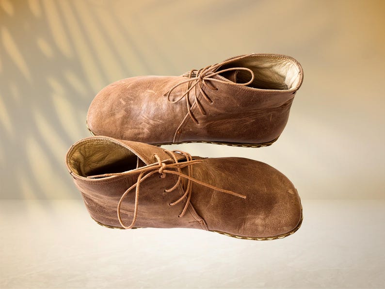 Puede incluir: Un par de botines de cuero marr&oacute;n con cordones. Las botas tienen un aspecto desgastado y texturizado y se colocan sobre un fondo neutro con una sombra sutil.