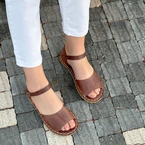 Pu&ograve; includere: Sandali in pelle marrone dal design semplice. I sandali hanno un cinturino alla caviglia e sono cuciti con filo bianco. I sandali sono aperti e sembrano fatti a mano.