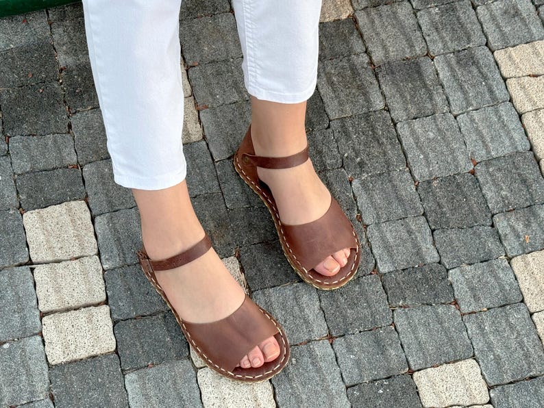 Pu&ograve; includere: Sandali in pelle marrone dal design semplice. I sandali hanno un cinturino alla caviglia e sono cuciti con filo bianco. I sandali sono aperti e sembrano fatti a mano.