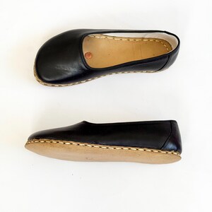 May include: Two black leather slippers with a rounded toe and a tan sole are shown against a white backdrop. The slippers have a simple design with visible stitching around the edges. The leather appears smooth and shiny.