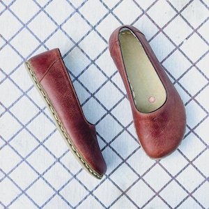May include: A pair of burgundy leather ballet flats. The shoes are displayed on a white surface with a blue grid pattern. The flats have a rounded toe and a stitched sole.