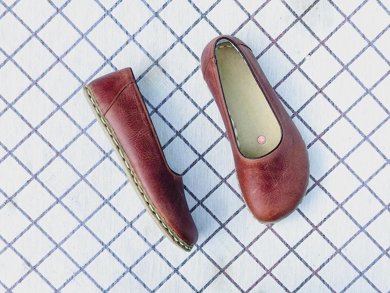 May include: A pair of burgundy leather ballet flats. The shoes are displayed on a white surface with a blue grid pattern. The flats have a rounded toe and a stitched sole.
