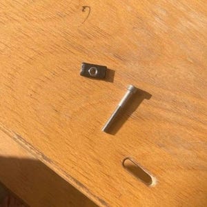 Könnte beinhalten: Eine silberne Schraube und ein schwarzes Metallstück mit einem Loch in der Mitte, beides liegt auf einer Holzoberfläche.