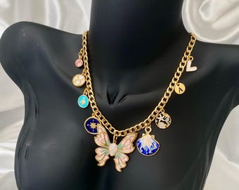 May include: Gold chain necklace with multiple charms, including a pink and white butterfly, a blue and white seashell, and a blue and white star charm. The necklace also features a pink, blue, and turquoise charm.
