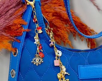 May include: A blue quilted handbag with a gold chain and charms. The charms include a heart, an apple, a crown, an evil eye, a perfume bottle, a bird, and an angel.