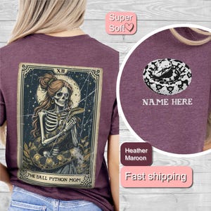May include: A maroon t-shirt with a graphic of a skeleton woman holding a snake. The graphic is in the style of a tarot card and says "The Ball Python Mom" at the bottom. The text "Super Soft" is in a pink box at the top of the image. The text "NAME HERE" is in a white box at the bottom of the image.