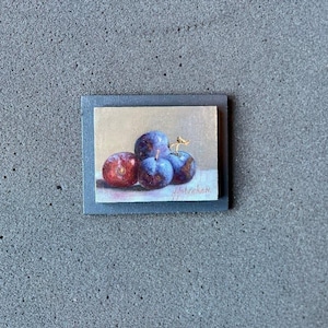 Könnte beinhalten: Ein kleines rechteckiges Gemälde mit vier Pflaumen in tiefen Violett-, Blau- und Rottönen vor einem neutralen Hintergrund. Das Kunstwerk ist auf einem dunkelgrauen Rahmen montiert und trägt eine Signatur in der unteren rechten Ecke.