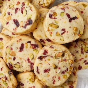 May include: A close-up of a pile of shortbread cookies with pistachios and dried cranberries. The cookies are light brown and have a slightly crispy texture.