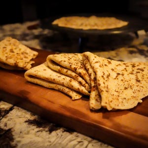 May include: A stack of six golden brown flatbreads, likely made from a simple dough, are arranged on a wooden cutting board. The flatbreads have a slightly crispy texture and are slightly browned on the surface.