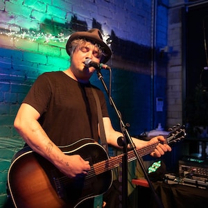 Könnte beinhalten: Ein Mann mit einem schwarzen Hut und einem schwarzen Hemd singt in ein Mikrofon, während er eine Akustikgitarre spielt. Die Gitarre ist braun mit einem schwarzen Schlagbrett. Der Mann steht vor einer blauen Backsteinwand mit grüner und violetter Beleuchtung.