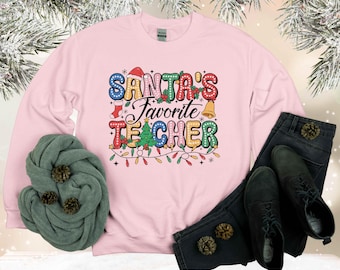Könnte beinhalten: Ein hellrosa Sweatshirt mit dem Text "Santa's Favorite Teacher" in bunten Buchstaben. Das Design umfasst einen Weihnachtsmannhut, einen Strumpf, eine Glocke, einen Weihnachtsbaum und Lichterketten. Das Sweatshirt wird mit schwarzen Jeans, Schuhen und einem grünen Schal kombiniert.