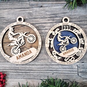 May include: Two wooden ornaments with a motocross theme. One is natural wood, the other has a blue center. Each features a rider on a motorcycle, with the name "Amanda" and "Amadeus" respectively. The ornaments have the words "Whip", "Air Time", and "Kickout".