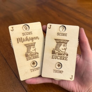 May include: Two wooden Euchre score cards are held in a hand. The cards are engraved with the letter "J", the word "SCORE", and the word "TRUMP". One card says "Michigan" and has a score of 9. The other card has a score of 7.