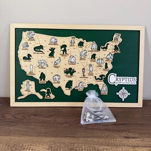 May include: A wooden map of the United States with cryptid illustrations, set against a green background within a wooden frame. A small bag of metal cryptid figurines sits in front of the map. The text "Cryptids of the united states" is visible.
