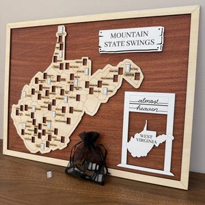 May include: A wooden wall map of West Virginia featuring cutouts for locations, a sign reading MOUNTAIN STATE SWINGS, and a white frame with the text almost heaven and WEST VIRGINIA. A black organza bag contains small wooden tokens for the map.
