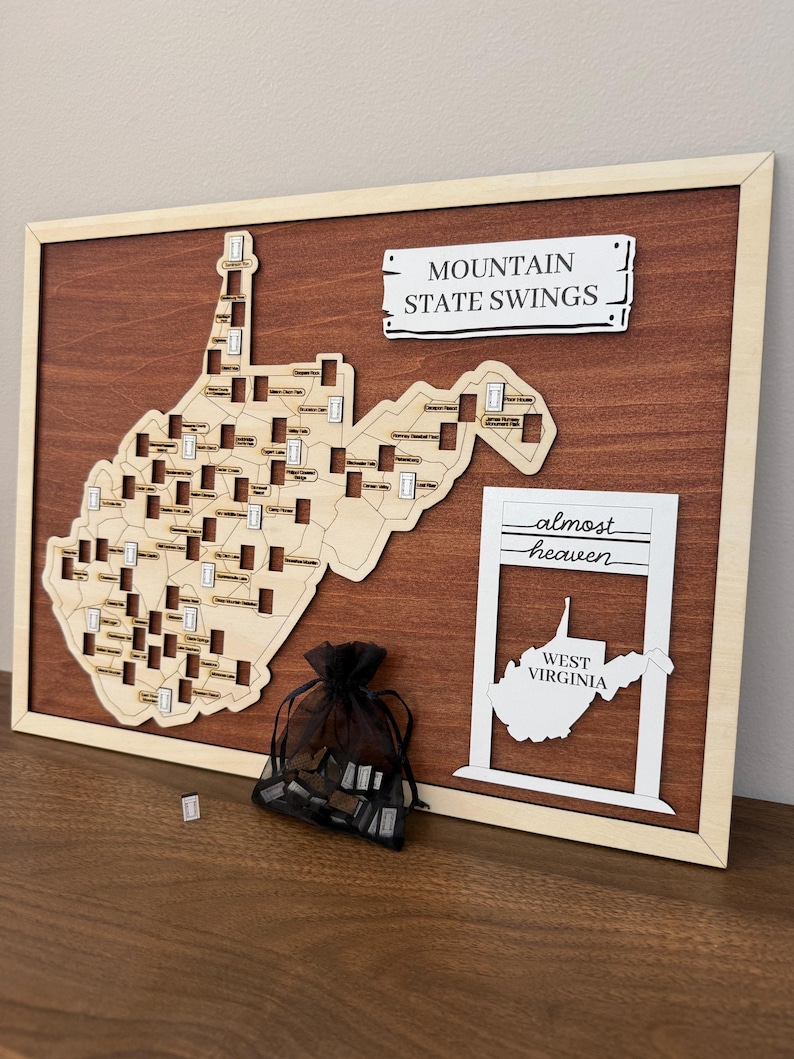 May include: A wooden wall map of West Virginia featuring cutouts for locations, a sign reading MOUNTAIN STATE SWINGS, and a white frame with the text almost heaven and WEST VIRGINIA. A black organza bag contains small wooden tokens for the map.